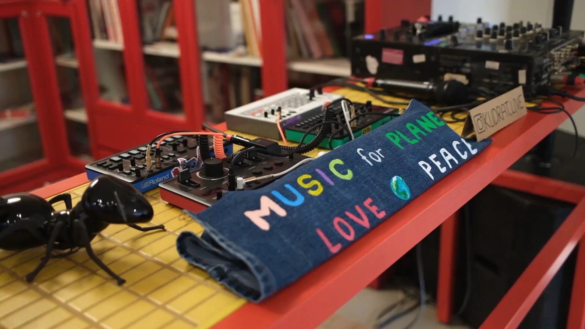 A hand-painted denim banner reading Music for Planet, Love, Peace, laid across a live rig of synths and drum machines